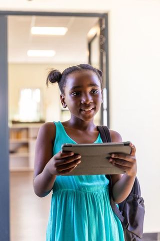 African american girl using tablet and standing in school doorway wearing turquoise dress