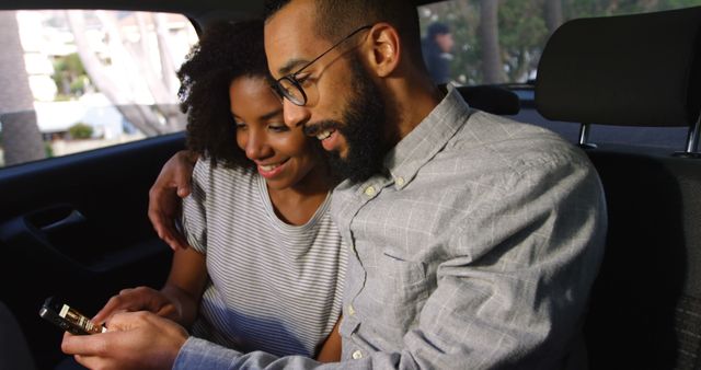 An African American couple sitting together in the backseat of a car, smiling and looking at a smartphone. Perfect for use in promoting technology, connectivity, and relationship-building moments. Great for apps, social media usage, online services, or automobile travel themes.