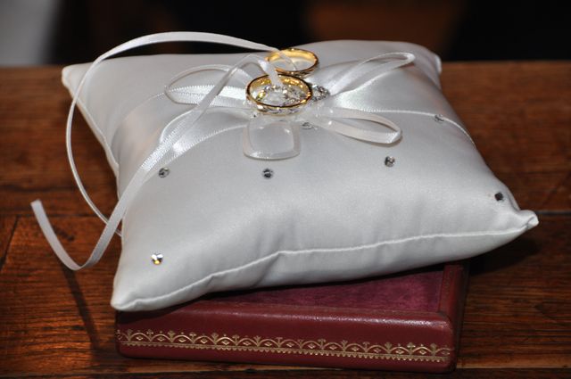 Photo depicts an elegant white cushion on a wooden table, adorned with wedding rings tied with a ribbon. Ideal for use in wedding invitations, ceremony decor catalogs, or event planning websites. Visualize the key moment of a wedding ceremony with a close-up view.