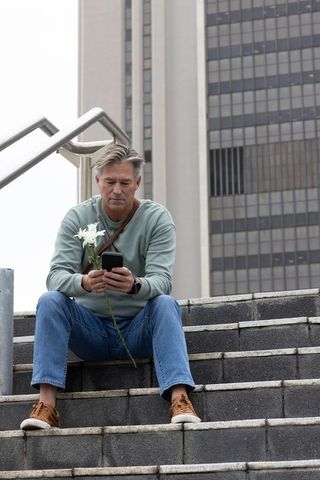 Middle-aged man sitting on city steps holding white flower and checking smartphone