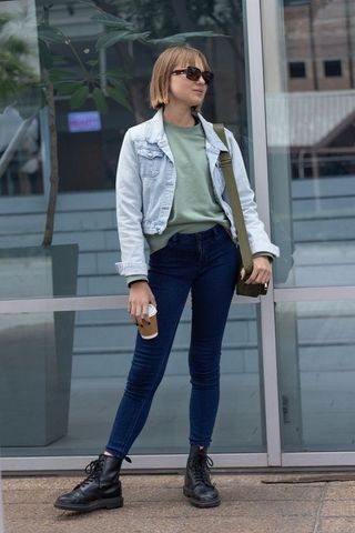 Young woman wearing denim jacket and sunglasses standing by glass facade holding coffee