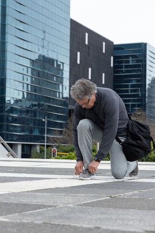 Senior man tying sneaker laces on geometric urban plaza with reflective glass towers