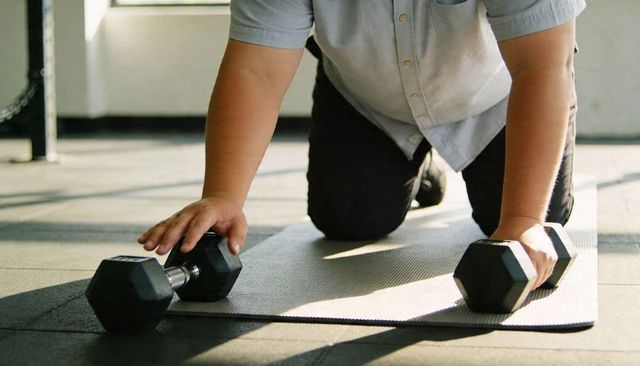 Kneeling man placing hands on hex dumbbells on mat in sunlit gym floor workout