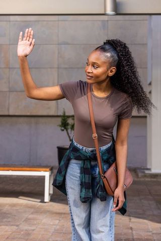 Young african american woman waving outdoors with crossbody bag and plaid tied at waist