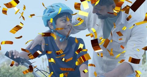 Father helping son ride bicycle while celebrating with golden confetti, family bonding