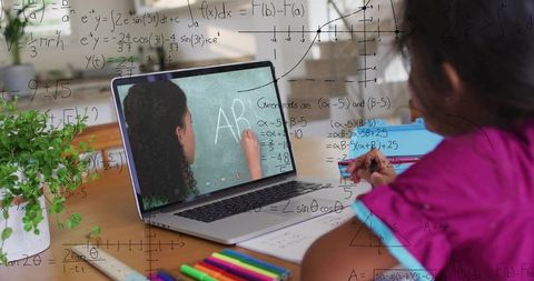 Teen studying math remotely on laptop while writing notes at wood desk with colorful pens