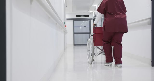 Healthcare worker wearing burgundy scrubs pushing a wheelchair down a clean, brightly lit hospital corridor. Ideal for use in content related to healthcare, nursing, hospital operations, patient care, clinical practice, and Medical support.