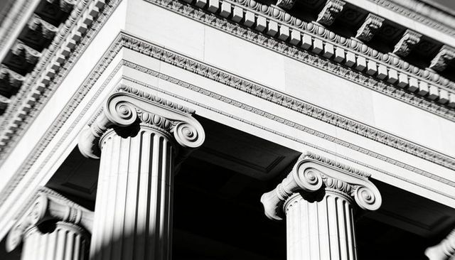 Low-angle view of three fluted ionic columns rising to ornate entablature in monochrome