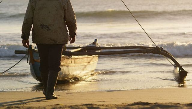 Fisherman walking with outrigger canoe on golden shoreline at sunrise for coastal lifestyle