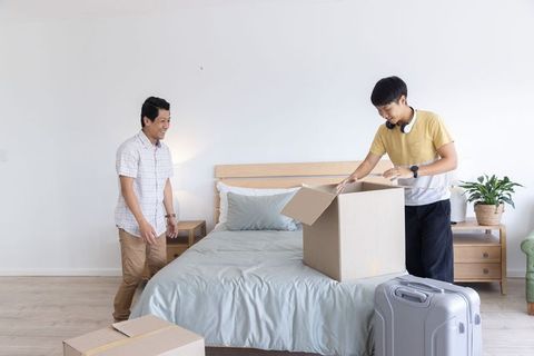 Two asian men unpacking boxes in bright minimal bedroom showing teamwork during relocation