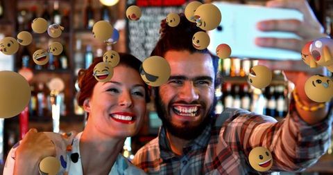 Friends Taking Selfie Surrounded by Floating Emojis in Lively Bar