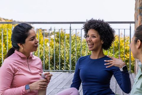 Diverse women chatting on deck in activewear, laughing and hydrating after workout outdoors