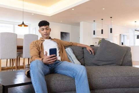 Young man lounging on sofa using smartphone in modern open-plan living room with clean decor