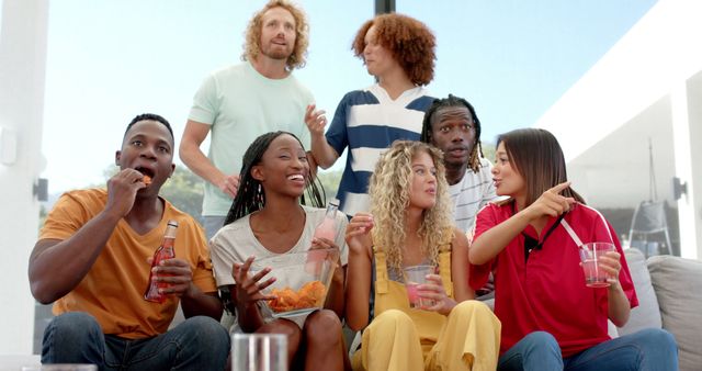 Diverse group of male and female colleagues watching tv and drinking and eating chips at home. Lifestyle, friendship, togetherness, entertainment, free time and domestic life, unaltered.