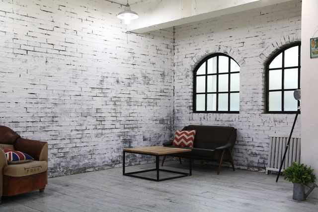 Modern industrial living room features whitewashed brick wall, arched windows, and minimalist furniture. Design includes a wooden table, vintage armchair, and plants creating a cozy and stylish interiorscape. Suitable for ideas on loft apartment designs, modern home décor, or renovations emphasizing simplicity mixed with industrial charm.