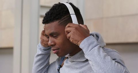 African american man adjusting white headphones wearing gray hoodie and backpack outdoors