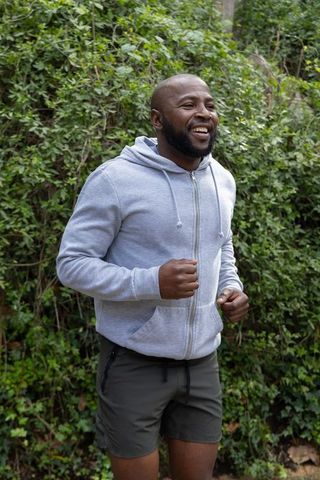 African american man jogging through leafy park wearing light gray zip-up hoodie and shorts