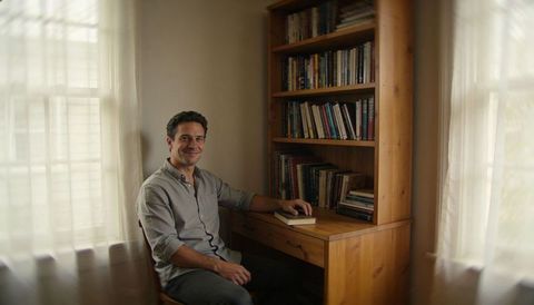Smiling man sitting at wooden desk in cozy home study with bookshelf and sheer curtains