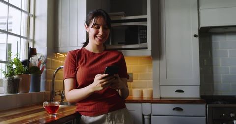 Smiling Woman in Kitchen Using Smartphone by Cup of Tea