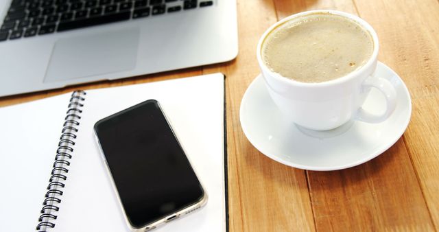 A cup of coffee, smartphone, and notepad rest on a wooden table next to a laptop, suggesting a work or study environment, with copy space. These items typically represent a modern professional or student setup, indicating productivity or a break.