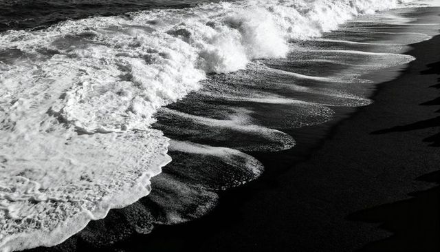 Monochrome surf creating scalloped foam patterns on black sand shoreline
