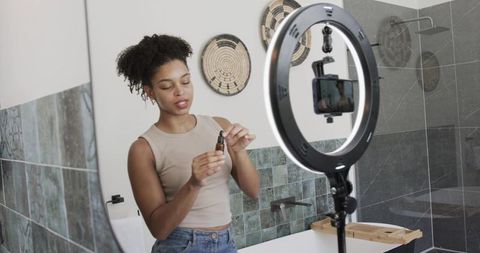 African woman filming skincare dropper with ring light in modern bathroom for beauty vlog