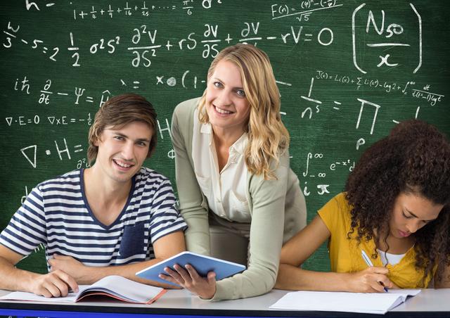 Smiling teacher assisting two students in a classroom with mathematical formulas on a green blackboard. Ideal for educational content, tutoring services, school websites, and academic resources.