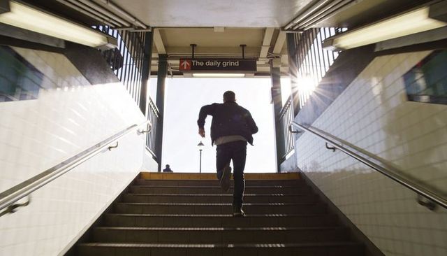 Commuter running up subway stairs toward daily grind sign with sunburst and lensflare