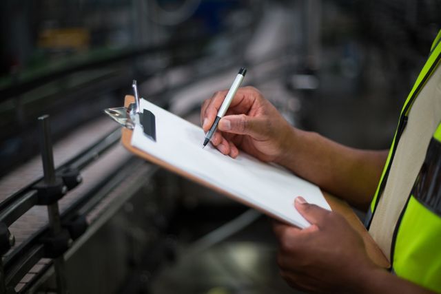 Mid section of factory worker writing on notepad in factory