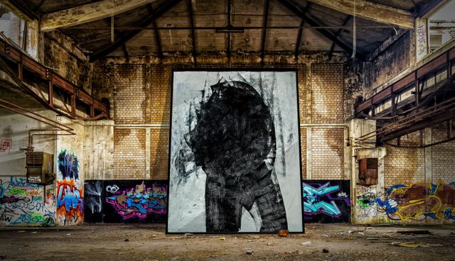 Abstract black and white portrait displayed on easel surrounded by vibrant graffiti inside deteriorating industrial building. Perfect usage for urban art themes, industrial decay visuals, or street art projects.