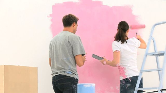 This visual captures a couple painting a white wall pink during a home renovation project. Utilizable for topics related to DIY home improvement, modern interior design, teamwork, and bonding activities for couples. Perfect for lifestyle blogs, instructional articles, and promotions for home improvement products or services.