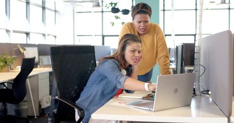 Diverse female coworkers collaborating at shared workstation in modern bright open office