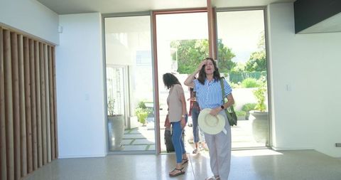 Sunlit modern foyer with visitors arriving, woman pausing with hat and shoulder bag