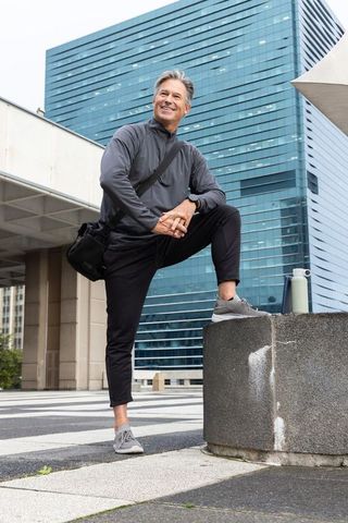 Smiling middle-aged man posing with foot on ledge in modern urban plaza with glass tower