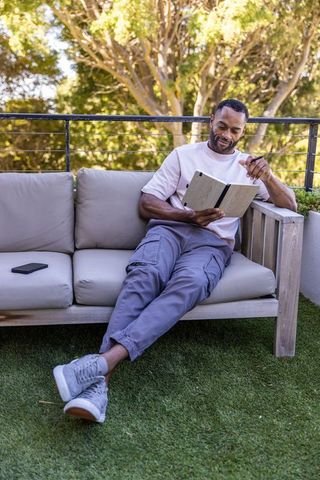 Mature african man relaxing on terrace sofa writing in notebook with pen and smartphone