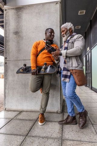 African american mentor and skateboarder checking smartphone while leaning on concrete pillar in urb