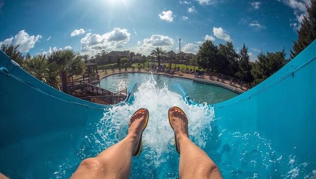 First-person sliding down blue water slide into resort pool, splashing legs in flip-flops