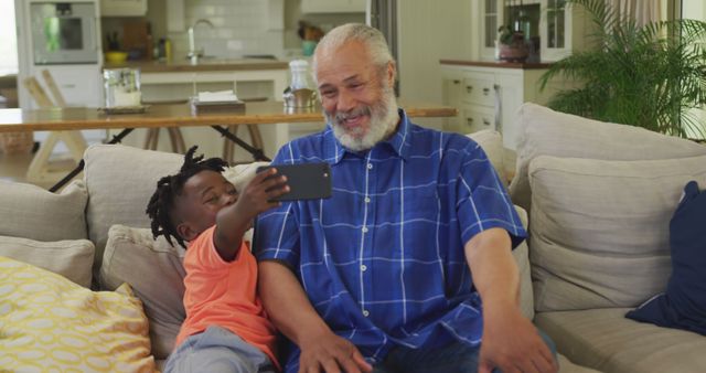 Elderly man with grey hair and beard sitting on couch with young grandson taking selfie using smartphone. Indoor living room space offers comfortable, relaxed atmosphere. Perfect for themes of family bonding, intergenerational relationships, modern technology use, happy moments, and home lifestyle.
