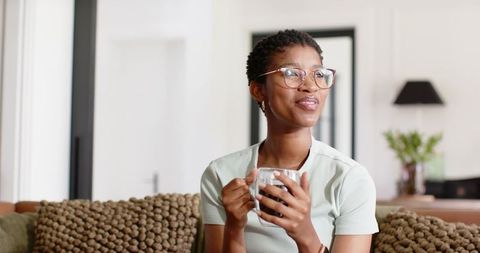African woman relaxing with coffee on chunky-knit sofa in bright modern cozy living room