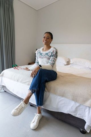Smiling african american woman sitting on bed in cozy knit sweater, denim and smartwatch