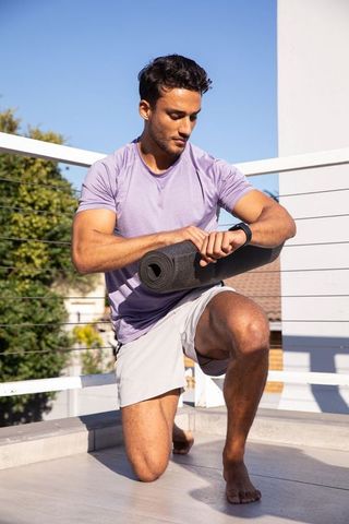 Fit man checking smartwatch while holding yoga mat on rooftop balcony