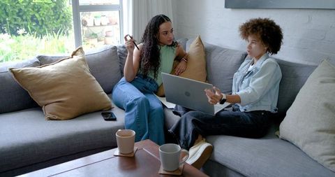 Two african american women discussing work on laptop while relaxing on gray sofa