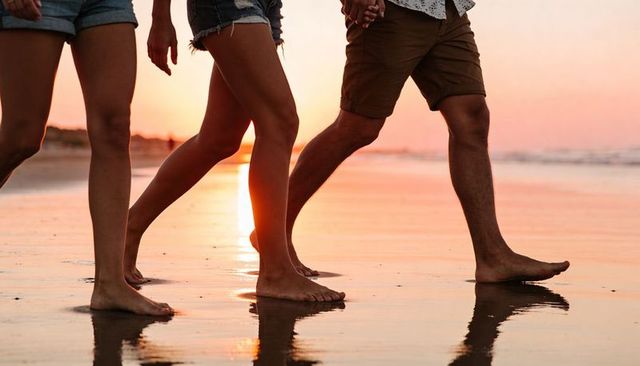 Three people walking barefoot on wet sand at sunset holding hands, romantic beach stroll