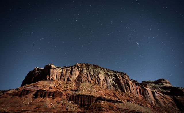 This nightscape features a dramatic view of a towering red rock desert cliff beneath a clear, star-filled sky. Ideal for use in outdoor adventure blogs, travel magazines, night sky enthusiasts' websites, geological study materials, or peaceful landscape themes showcasing natural wilderness and tranquility.