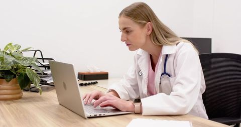 Female physician typing on laptop in clinic office with stethoscope and plant in modern workspace