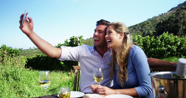 A young Caucasian couple is taking a selfie while enjoying a wine tasting experience outdoors, with copy space. Their cheerful expressions and the vineyard backdrop suggest a romantic and leisurely day spent together.