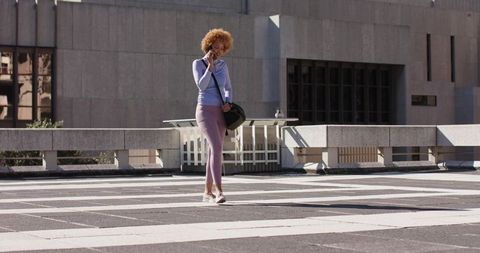 African American woman walking across urban crosswalk talking on phone carrying black bag