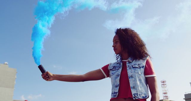 Young woman holding blue smoke bomb under clear sky, expressing confidence and freedom. Dressed in a red shirt and denim vest, she stands in an urban outdoors area, demonstrating independence and creativity. Ideal for campaigns focused on youth culture, outdoor activities, fashion, or celebrating individualism.