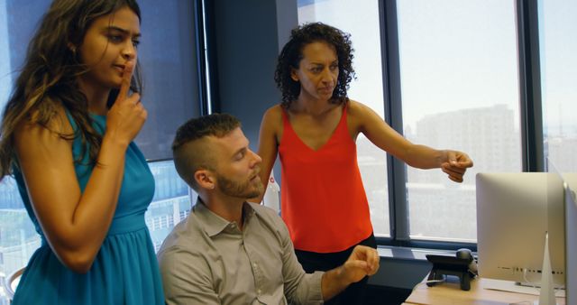 Three coworkers, representing different ethnic backgrounds, collaborating and discussing a project in a modern office space. One woman points at the computer screen while the other two concentrate on the details. Ideal for images representing teamwork, diversity in the workplace, collaborative efforts, and modern business environments.
