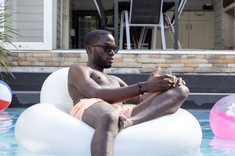 African american man relaxing on white inflatable ring in backyard pool with drink
